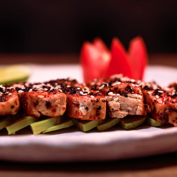 Grilled chicken skewers with sesame seeds and avocado slices, garnished with red tomato petals.