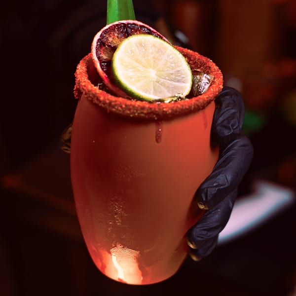 A hand in a black glove holding a red michelada with lime and orange slices.