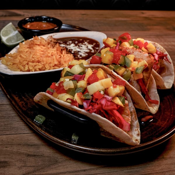 Three Clase Codigo tacos with colorful salsa, rice, beans, and a lime wedge on a dark plate.