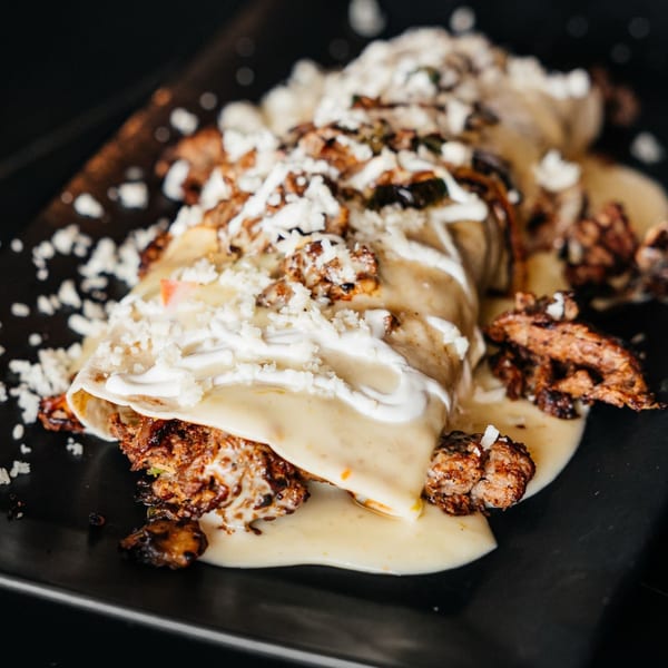 Mexican enchilada topped with creamy sauce, white cheese, and crumbled meat on a black plate.